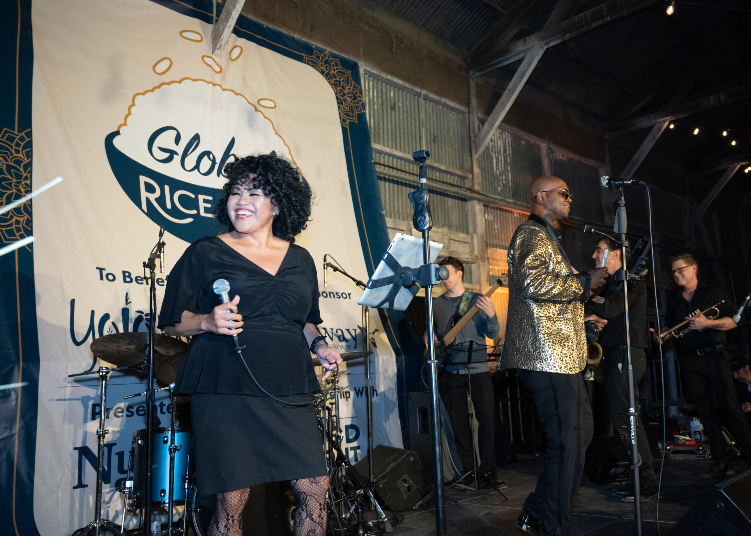 Woman singer smiling, man in gold jacket singing, and the band members in the background. All on stage